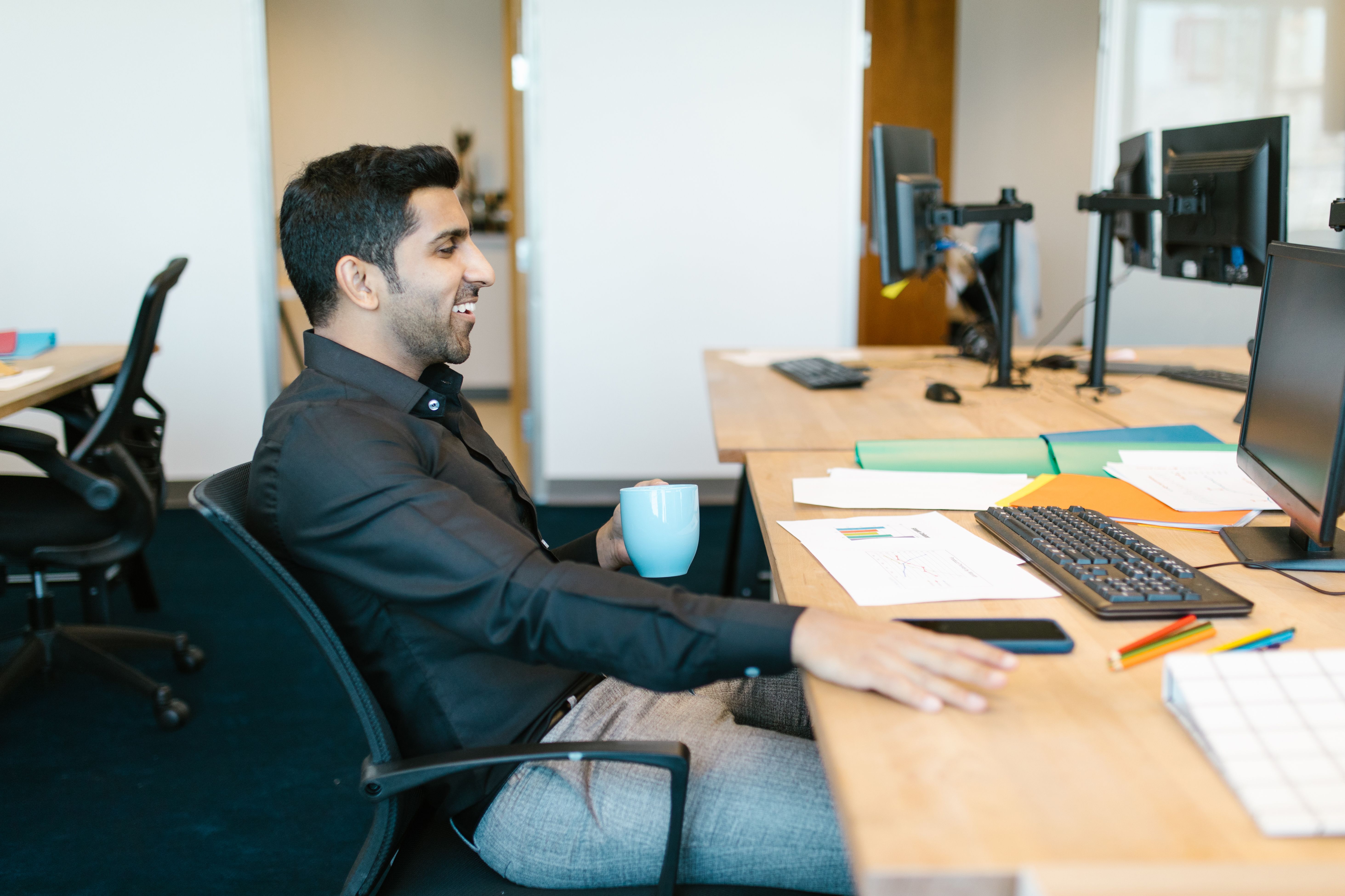 A man in a black shirt sits at a desk with computer monitors, holding a blue mug and smiling, surrounded by papers and stationery.