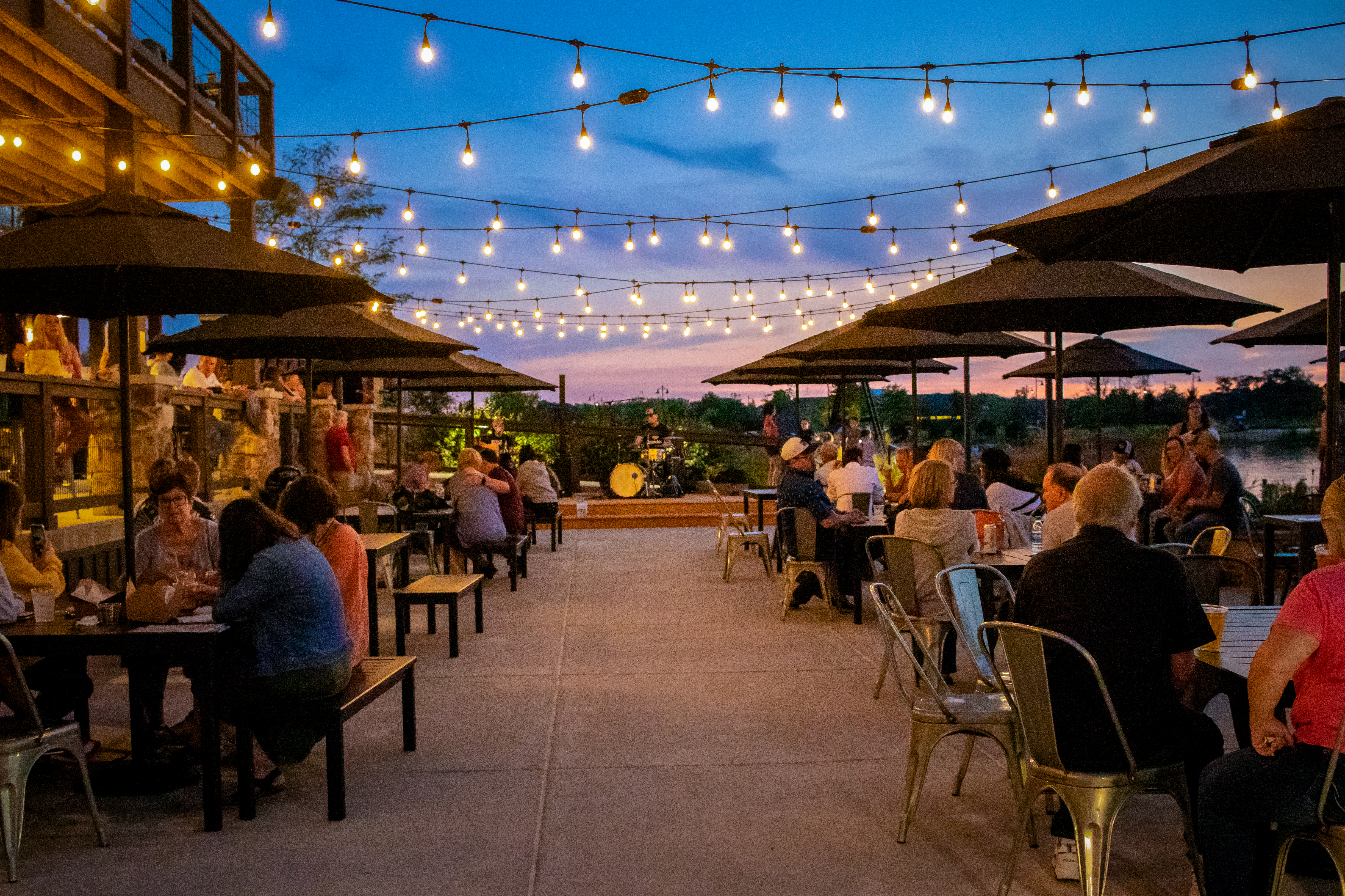 Outdoor patio dining area at sunset with string lights overhead, people seated at tables under umbrellas, and a band performing in the background.