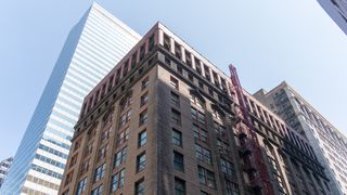 View of two contrasting buildings, one modern with glass panels, the other historic with brick facade and red fire escape, under a clear blue sky.