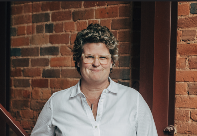 A person with curly hair and glasses, wearing a white shirt, smiles lightly while standing by a brick wall.