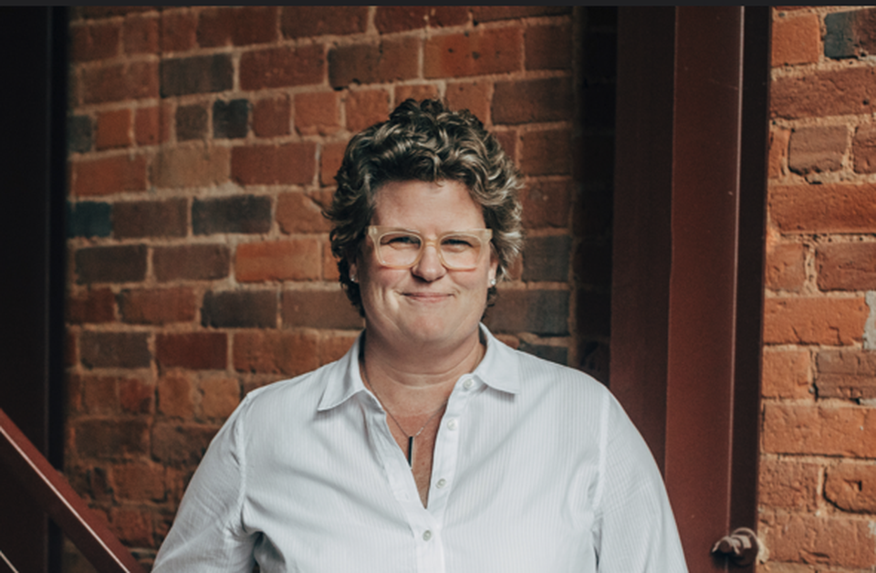 A person with curly hair and glasses, wearing a white shirt, smiles lightly while standing by a brick wall.