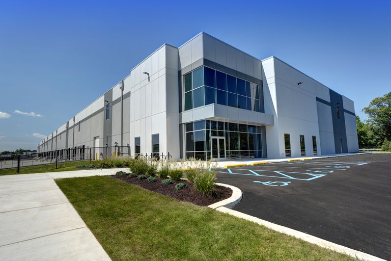 Modern industrial building with large glass windows and a landscaped exterior, adjacent to a paved parking area under a clear blue sky.