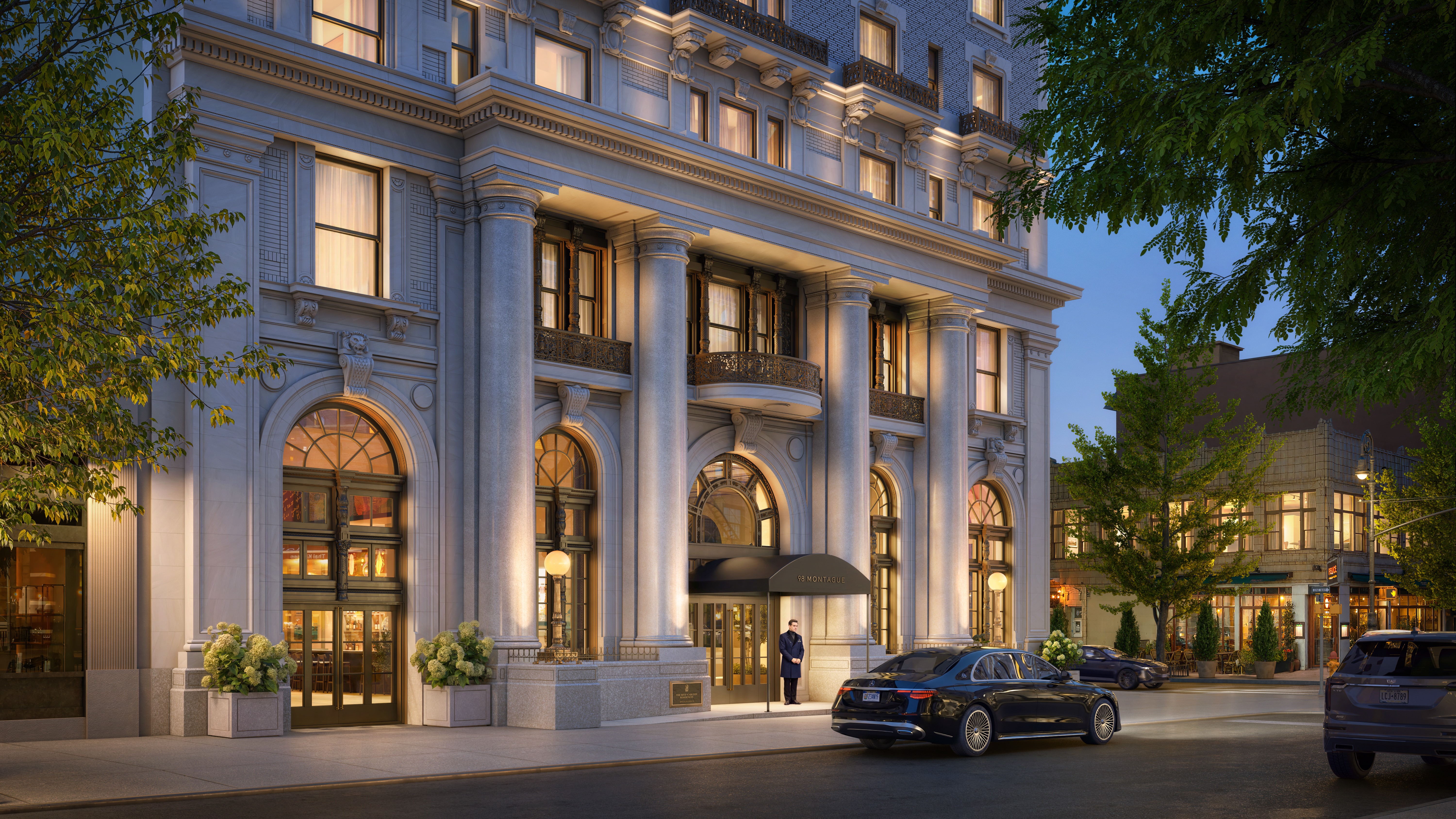 Elegant multi-story building with large columns, arched windows, and warm lighting. A doorman stands at the entrance with parked cars nearby.
