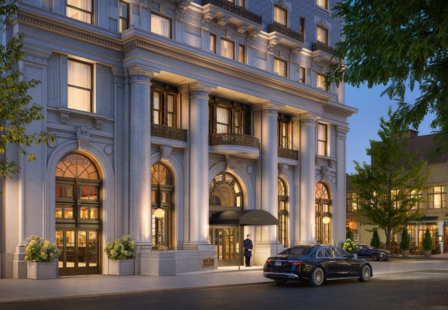 Elegant multi-story building with large columns, arched windows, and warm lighting. A doorman stands at the entrance with parked cars nearby.