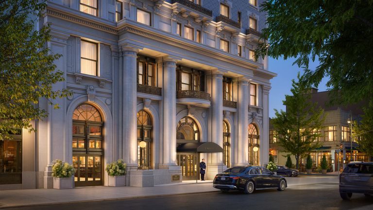Elegant multi-story building with large columns, arched windows, and warm lighting. A doorman stands at the entrance with parked cars nearby.
