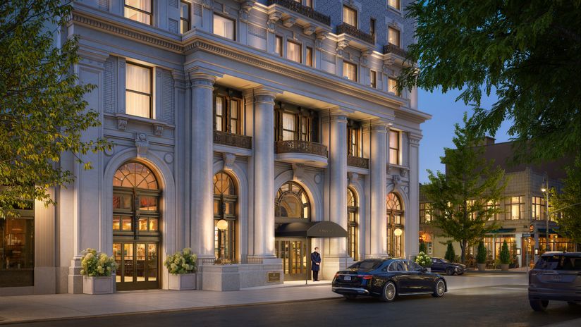 Elegant multi-story building with large columns, arched windows, and warm lighting. A doorman stands at the entrance with parked cars nearby.