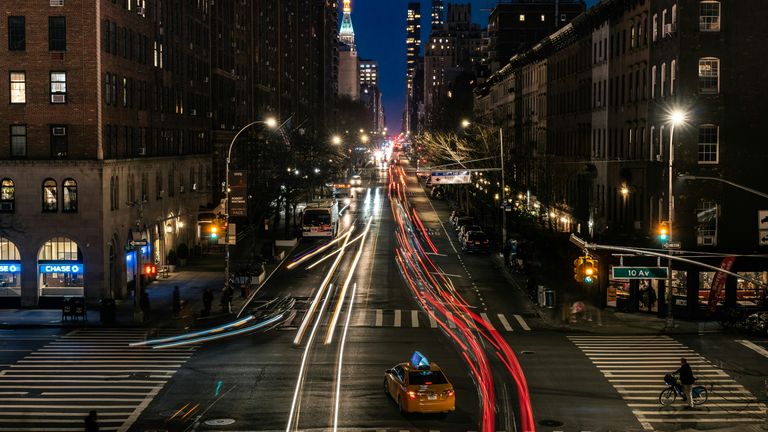City street at night with light trails from vehicles, a yellow taxi turning, bright storefronts, and a cyclist at the intersection.
