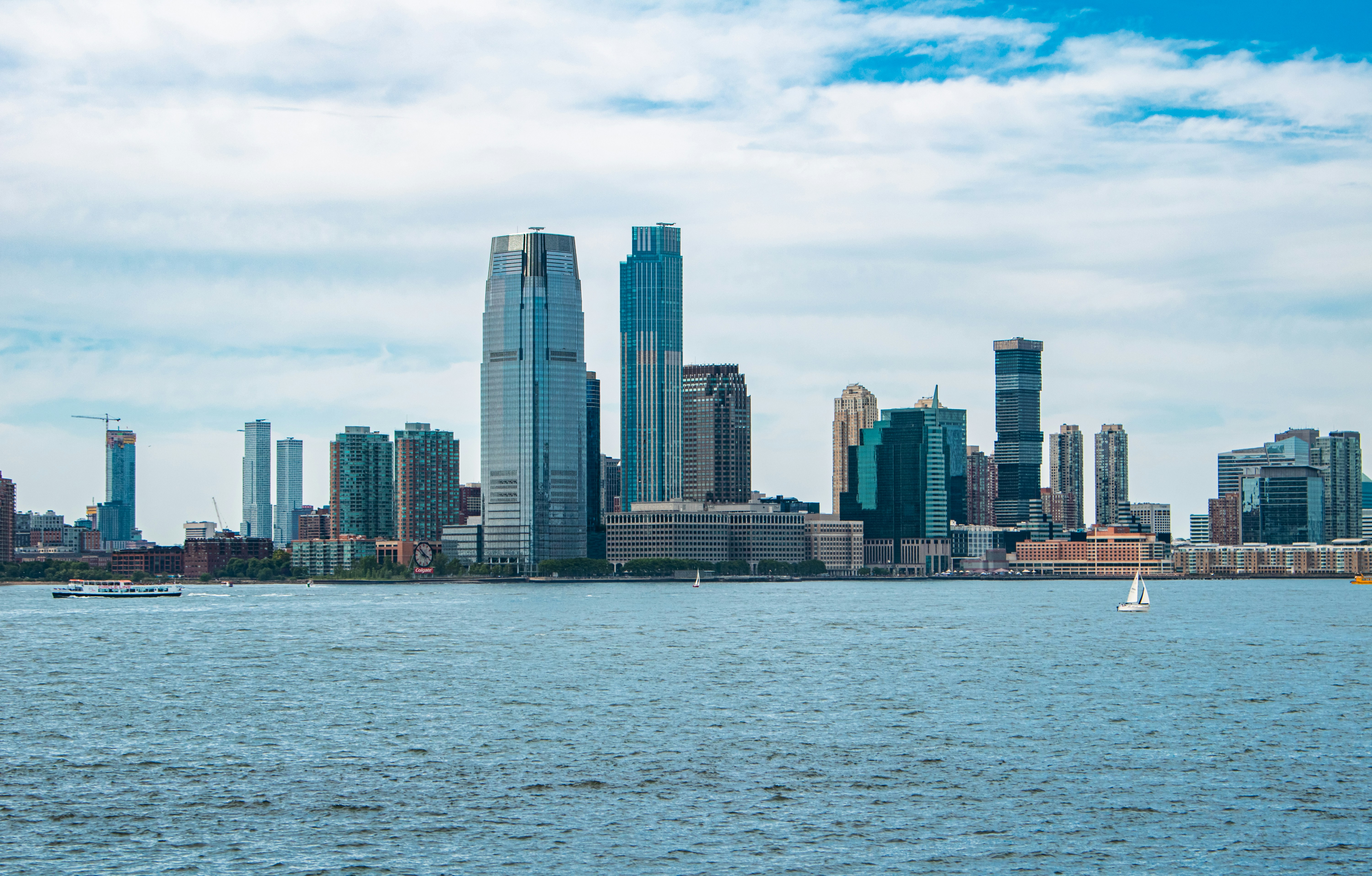 Skyline of a modern city with tall skyscrapers near a river, under a partly cloudy blue sky. A boat and sailboat are on the water.
