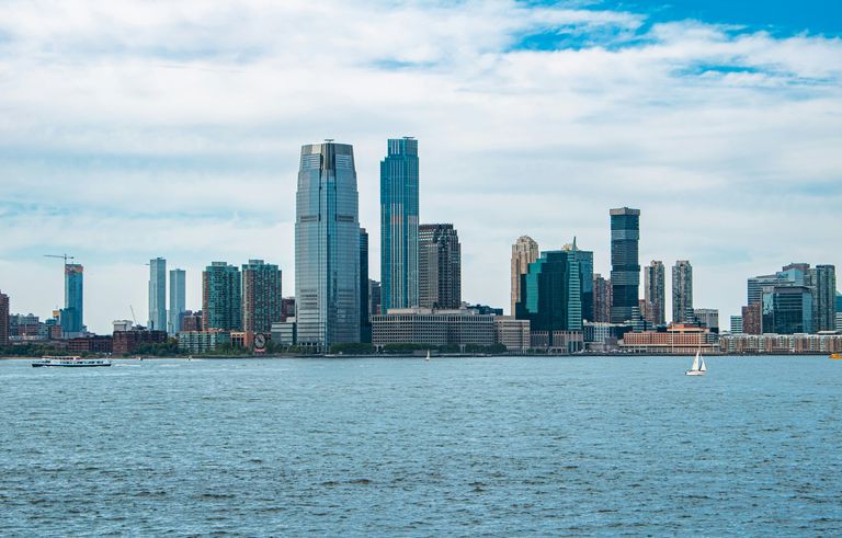 Skyline of a modern city with tall skyscrapers near a river, under a partly cloudy blue sky. A boat and sailboat are on the water.