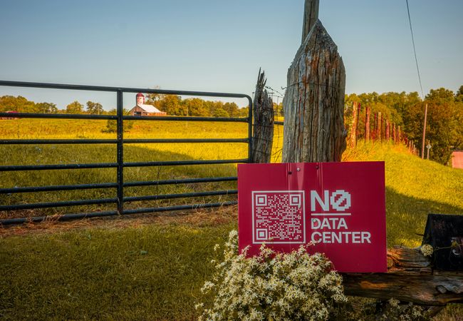 A red sign with "No Data Center" and a QR code is mounted on a gatepost by a field with a distant barn under a clear sky.