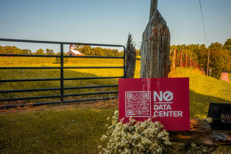 A red sign with "No Data Center" and a QR code is mounted on a gatepost by a field with a distant barn under a clear sky.