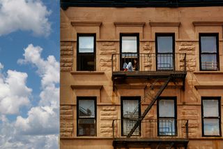 A person sitting on a fire escape of a brownstone building against a backdrop of a bright blue sky with white clouds.