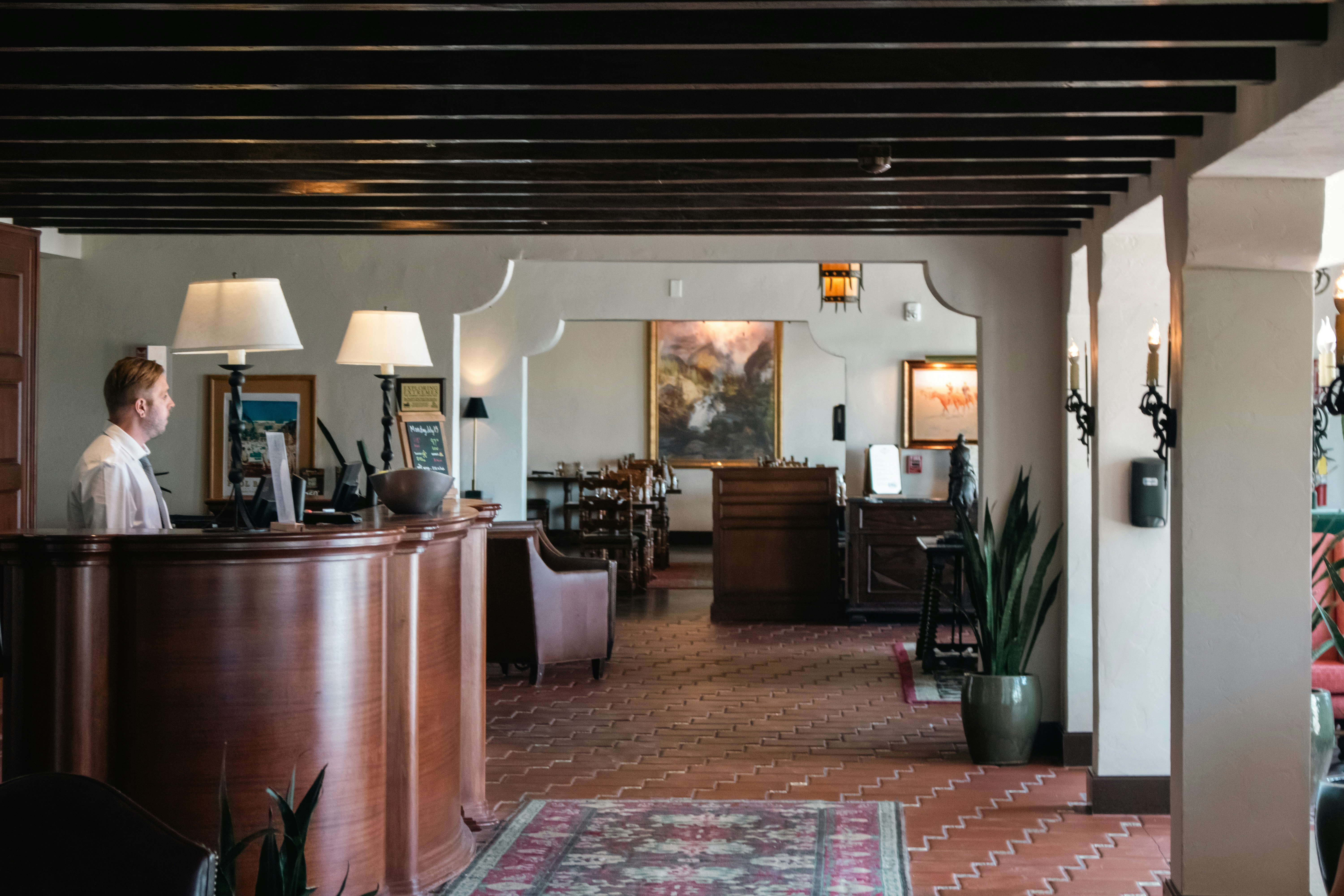 A lobby with dark wood beams, a concierge desk, and warm lighting. There's a person standing at the desk, and seating with artwork on the walls.
