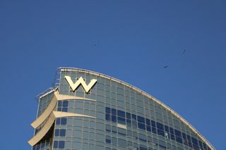 Glass skyscraper with a large white "W" on top, set against a clear blue sky, with a few birds flying nearby.