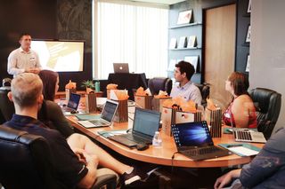 A group of people sit around a conference table with laptops and gift bags, attentively listening to a presenter in a modern office setting.
