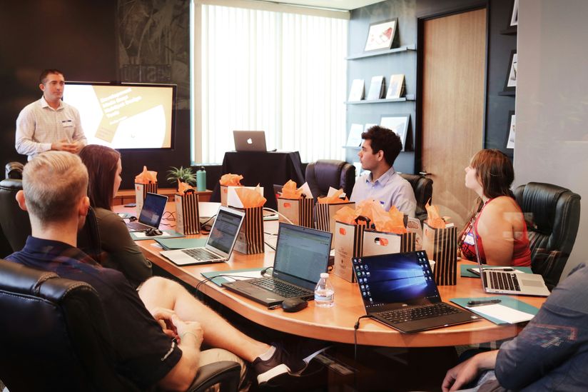 A group of people sit around a conference table with laptops and gift bags, attentively listening to a presenter in a modern office setting.