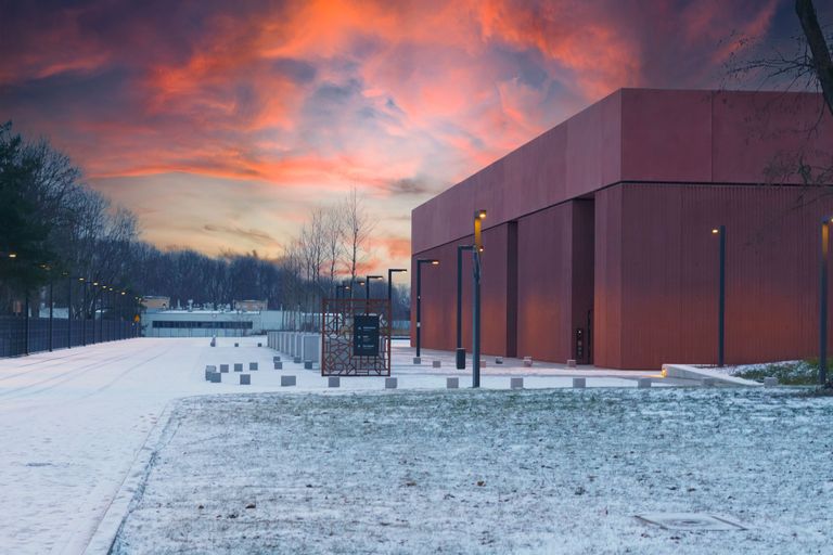 Modern red building at sunset with snow on the ground, surrounded by streetlights and trees in the background.