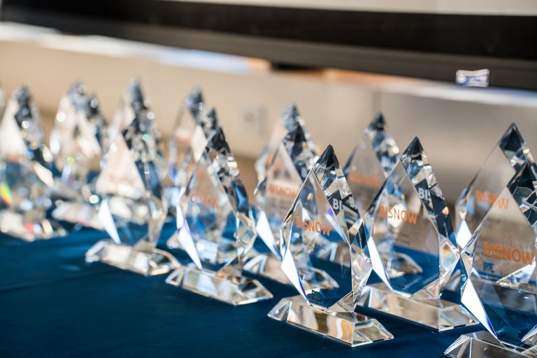 A row of clear crystal awards with "BISNOW" inscription on a blue tablecloth.