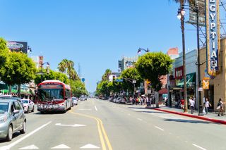 A busy urban street with a bus, cars, palm trees, shops, and pedestrians under a clear blue sky.