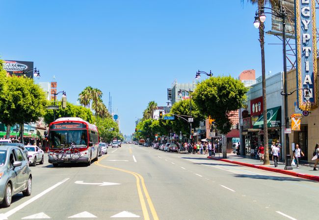 A busy urban street with a bus, cars, palm trees, shops, and pedestrians under a clear blue sky.