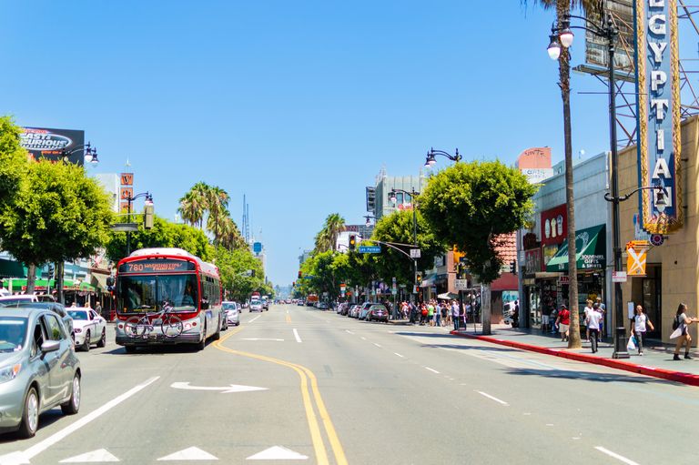 A busy urban street with a bus, cars, palm trees, shops, and pedestrians under a clear blue sky.