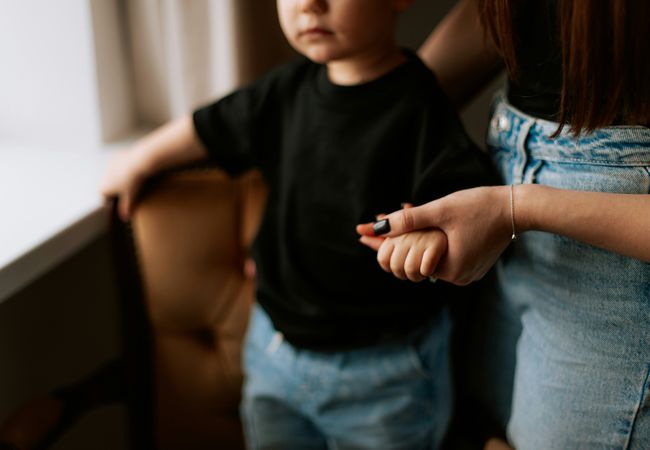 Woman holding a child's hand, both wearing casual clothes. The child is wearing a black shirt and blue jeans, standing by a window.