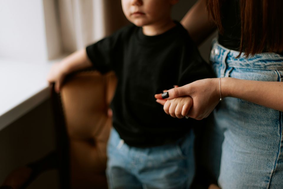Woman holding a child's hand, both wearing casual clothes. The child is wearing a black shirt and blue jeans, standing by a window.