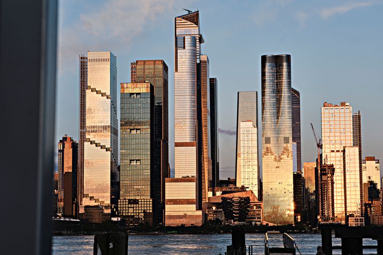 City skyline with modern skyscrapers reflecting the warm hues of sunset over a river, with a crane visible in the background.