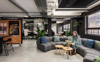 Modern office space with gray sectional sofa, plants, and people working. A woman uses a laptop on the couch, and others work at desks.