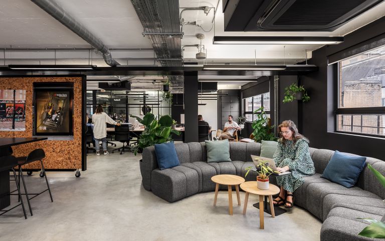 Modern office space with gray sectional sofa, plants, and people working. A woman uses a laptop on the couch, and others work at desks.
