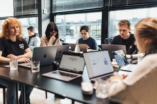 A group of people working on laptops at a table in a modern office setting, with focused expressions and natural light from large windows.