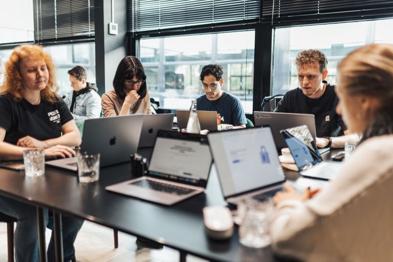 A group of people working on laptops at a table in a modern office setting, with focused expressions and natural light from large windows.