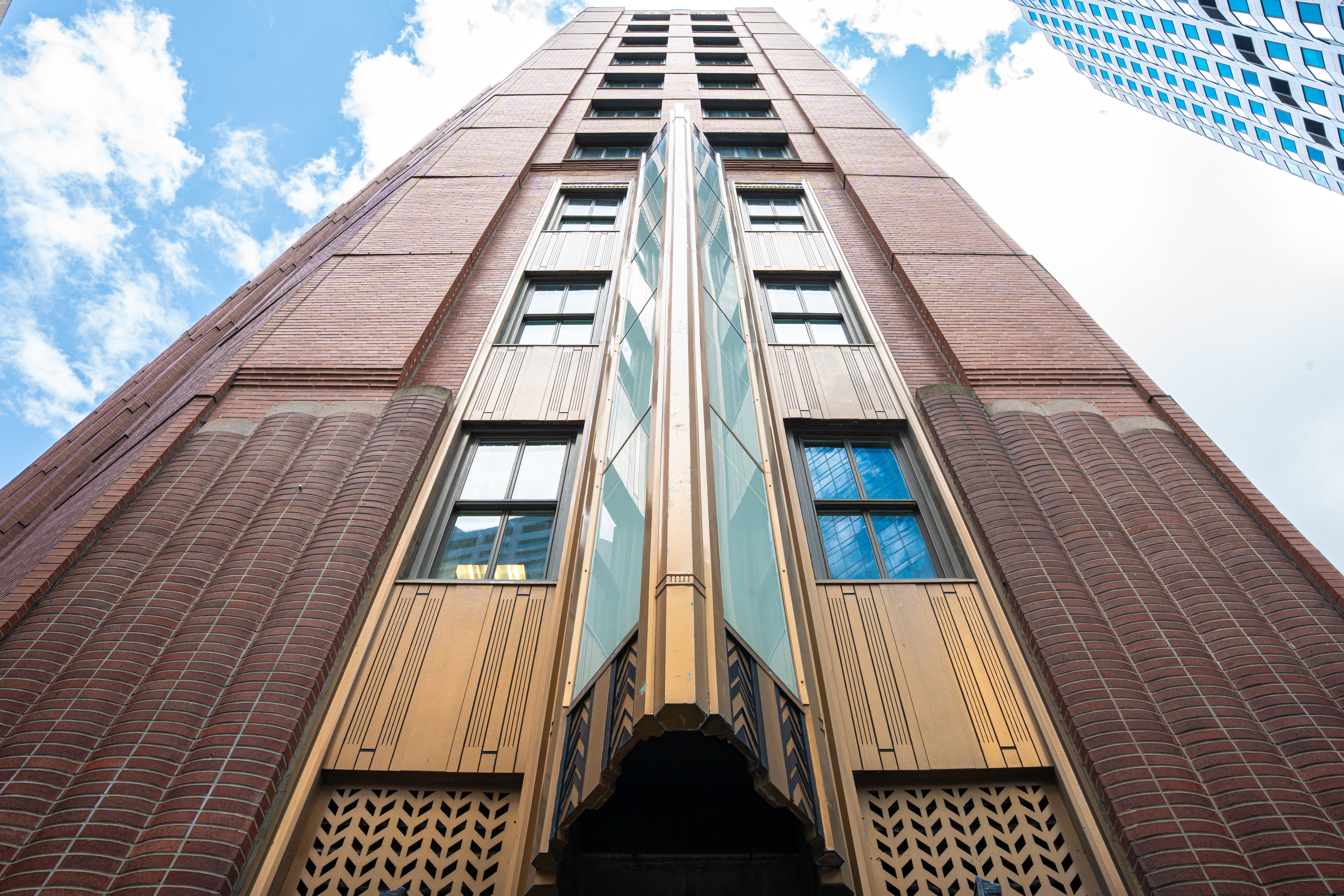 Upward view of a tall brick building with art deco design elements, featuring decorative patterns and large windows against a blue sky with clouds.