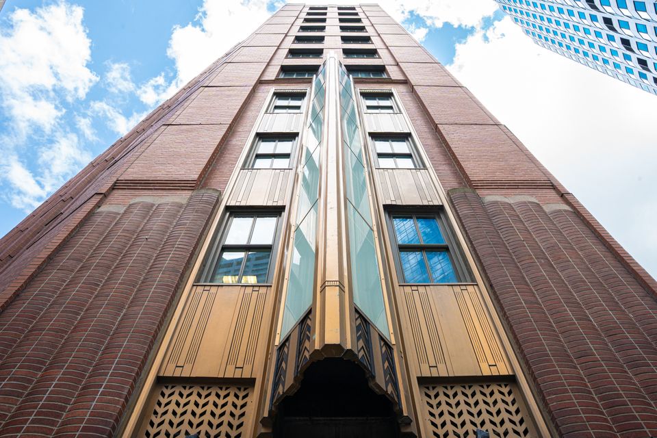 Upward view of a tall brick building with art deco design elements, featuring decorative patterns and large windows against a blue sky with clouds.