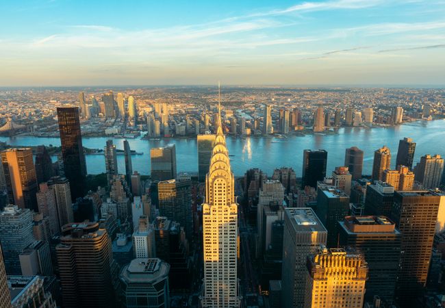 Aerial view of New York City skyline featuring the Chrysler Building, with the East River and Queens in the background under a blue sky at sunset.
