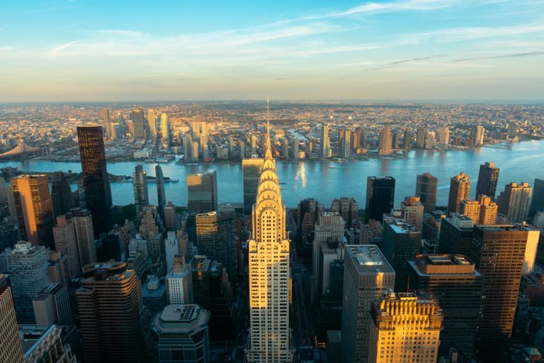 Aerial view of New York City skyline featuring the Chrysler Building, with the East River and Queens in the background under a blue sky at sunset.