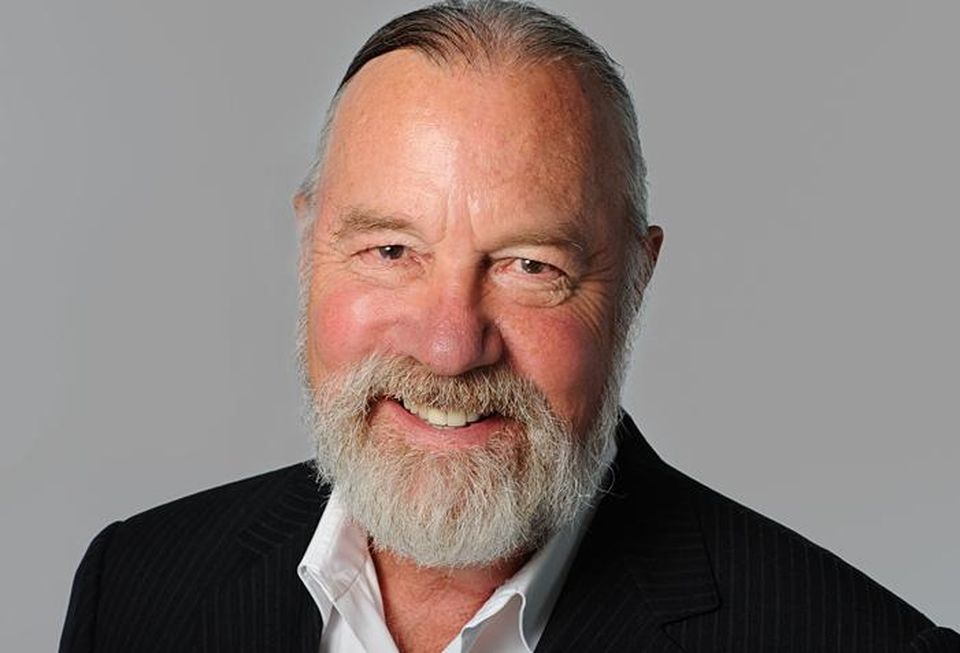 A professional headshot of Marcel Arsenault against a gray background. He's wearing a black blazer and white shirt with the top unbuttoned.