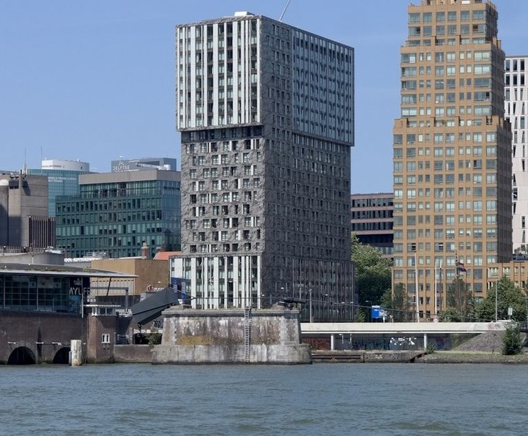 Modern high-rise buildings near a waterfront, with one uniquely designed structure featuring a square top, under a clear blue sky.