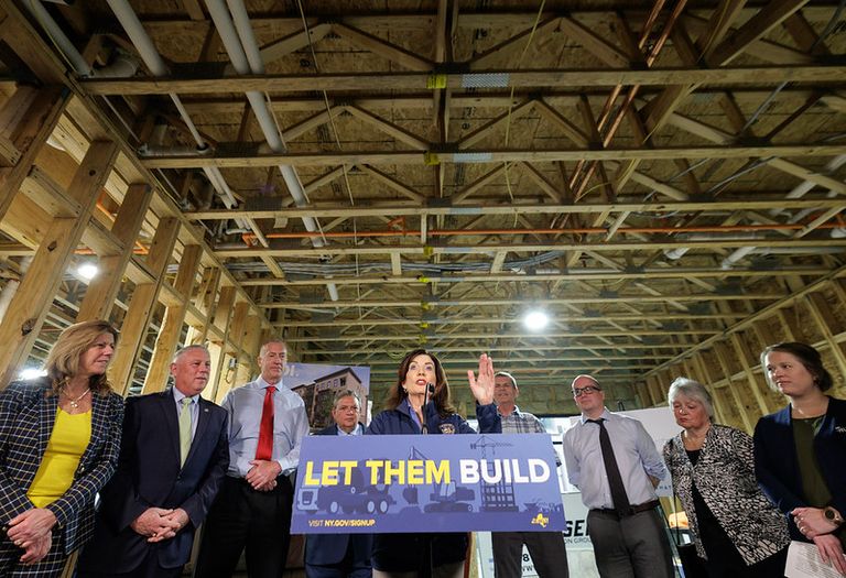 Gov. Kathy Hochul speaks at a podium with a "Let Them Build" sign, surrounded by several people inside a building under construction.