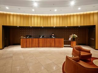 Hotel lobby with a wooden reception desk, circular wall decor, a staff member, leather chairs, and warm lighting and tones.