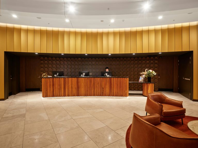 Hotel lobby with a wooden reception desk, circular wall decor, a staff member, leather chairs, and warm lighting and tones.