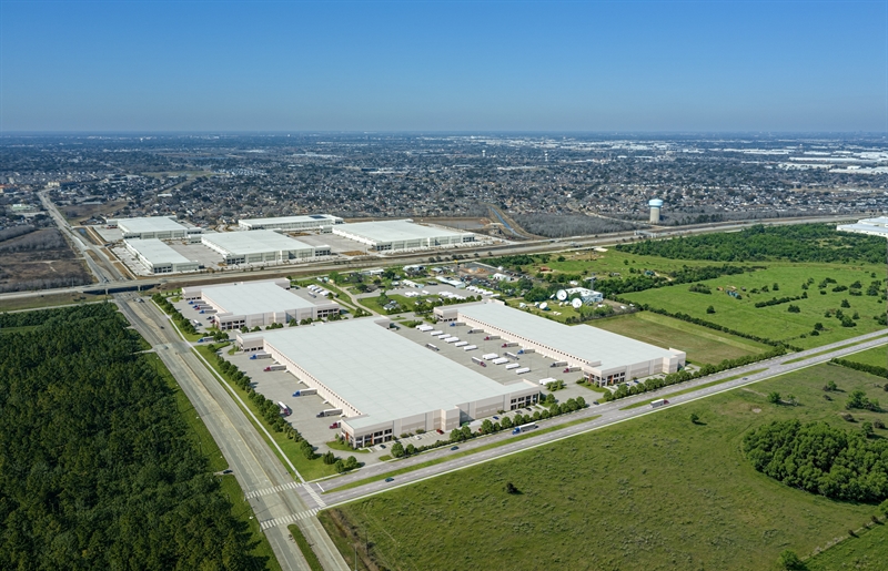 Aerial view of a large industrial park with warehouses surrounded by grass and trees, near a cityscape under a clear blue sky.