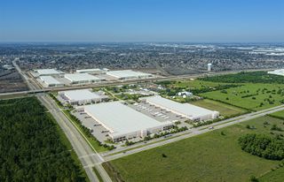 Aerial view of a large industrial park with warehouses surrounded by grass and trees, near a cityscape under a clear blue sky.