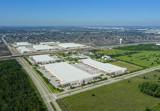Aerial view of a large industrial park with warehouses surrounded by grass and trees, near a cityscape under a clear blue sky.