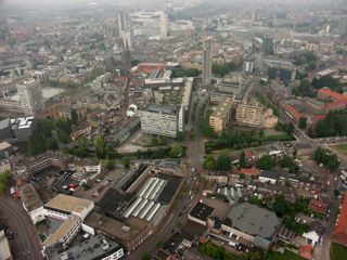 Aerial view of a city with a mix of modern and traditional architecture, featuring tall buildings, winding roads, and green spaces under a cloudy sky.