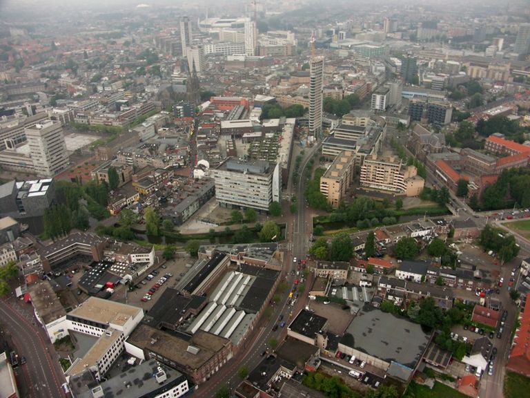 Aerial view of a city with a mix of modern and traditional architecture, featuring tall buildings, winding roads, and green spaces under a cloudy sky.