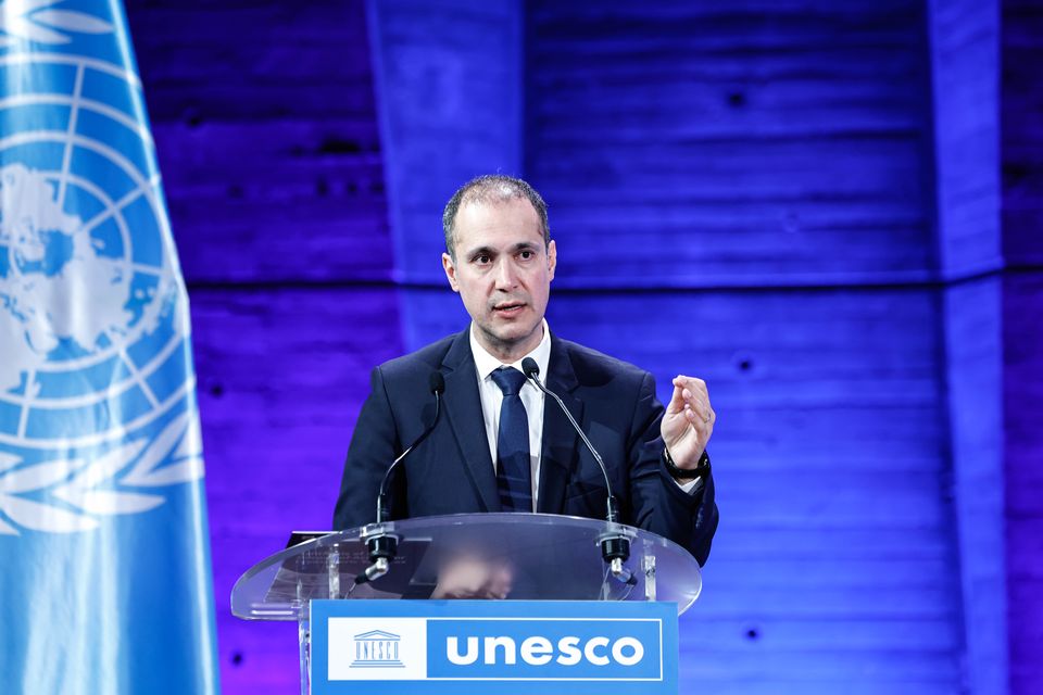 A man in a suit speaks at a podium with UNESCO branding, alongside a United Nations flag, set against a blue-lit background.