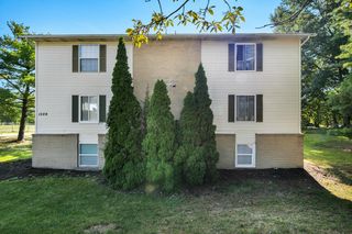 A two-story beige apartment building with dark shutters, a brick foundation, and tall evergreen shrubs in the front yard, set against a sunny, wooded backdrop.