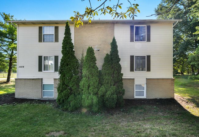 A two-story beige apartment building with dark shutters, a brick foundation, and tall evergreen shrubs in the front yard, set against a sunny, wooded backdrop.