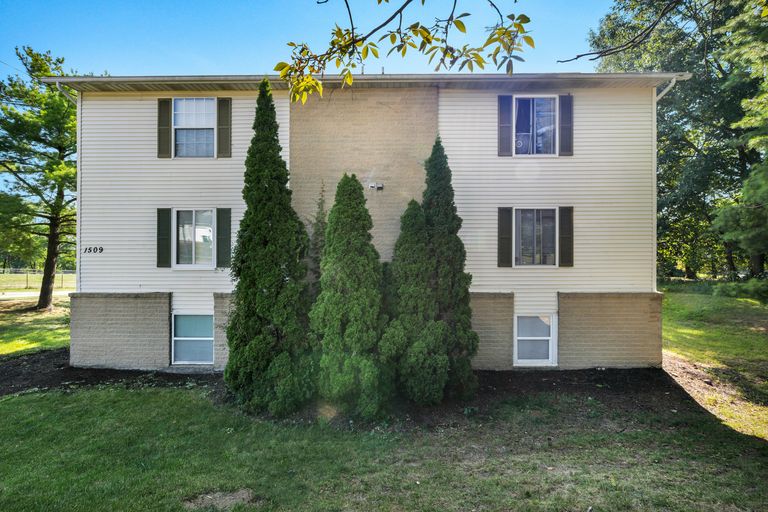 A two-story beige apartment building with dark shutters, a brick foundation, and tall evergreen shrubs in the front yard, set against a sunny, wooded backdrop.